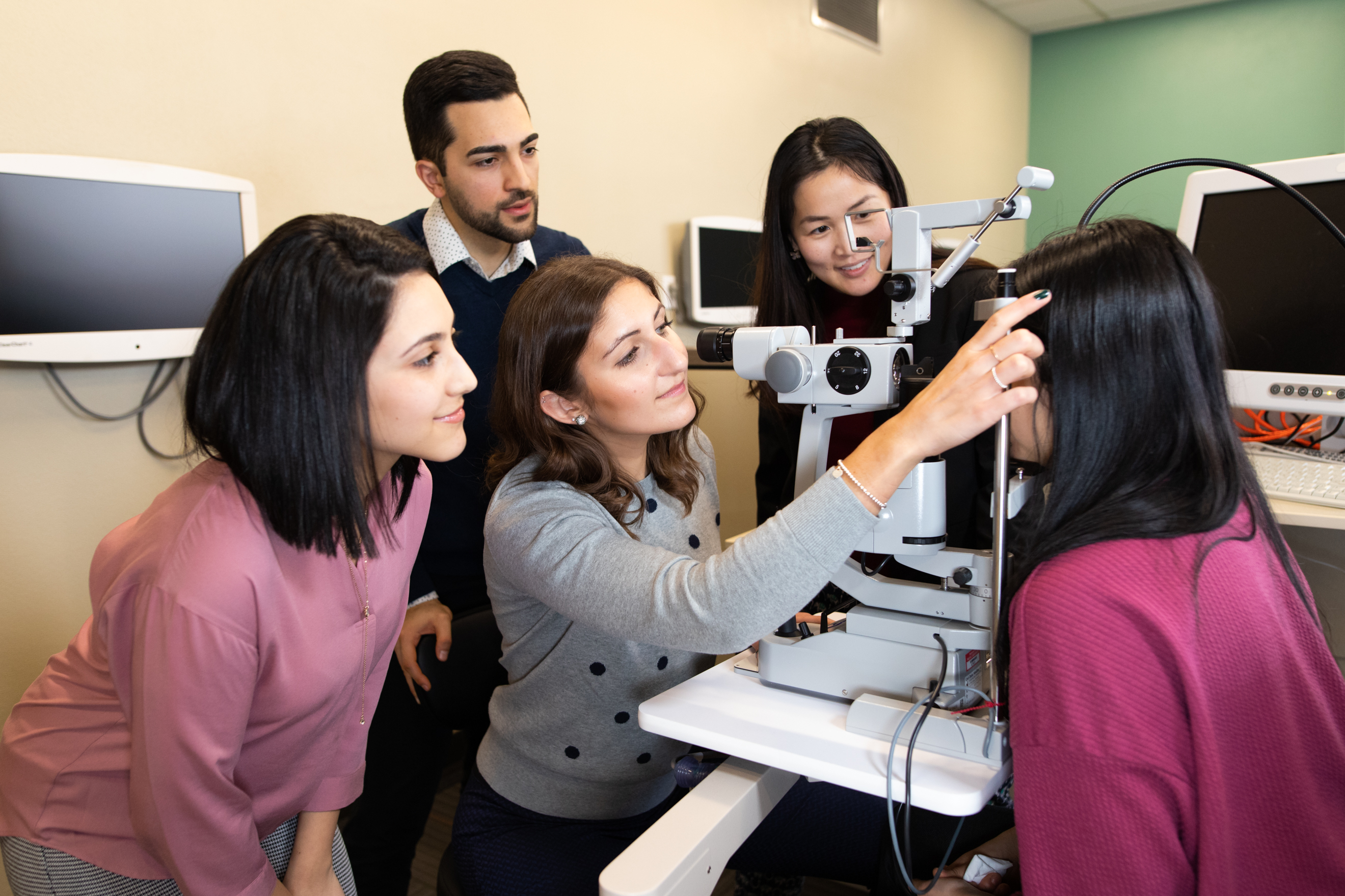 A group of student optometrists gather around a medical device as they examine a patient's eyes. 
