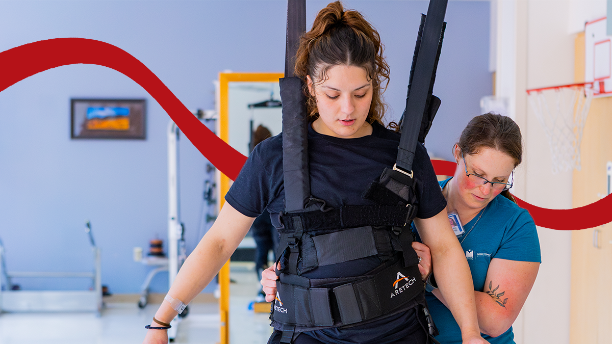 A Pacific alumna wearing blue scrubs helps a demonstration patient walk using a harness..