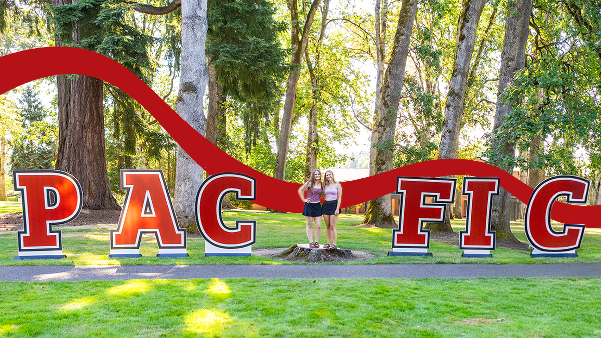 Two female students stand on a stump in place of the first "I" in a giant PACIFIC sign.