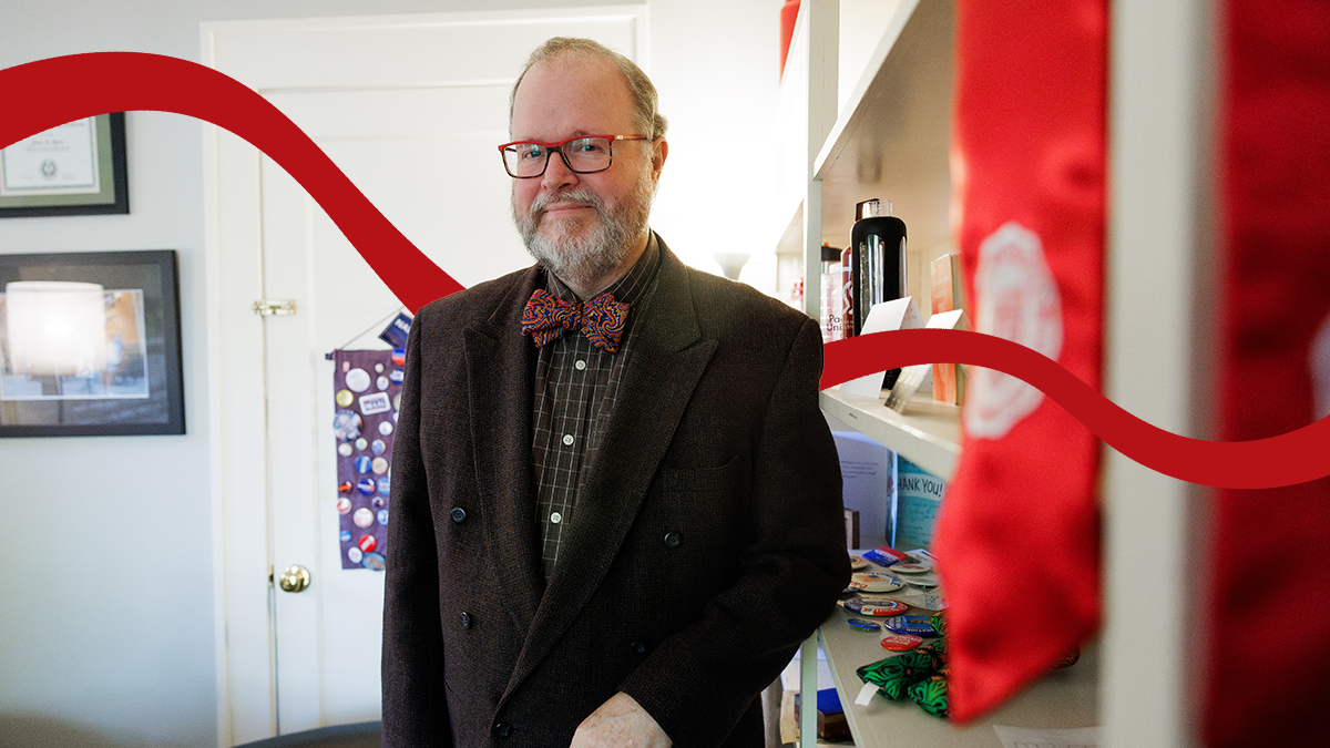 Pacific Professor Jim Moore stands in his office wearing a suit and bowtie. Beyhind him is a banner and shelves, both featuring a variety of campaign buttons.