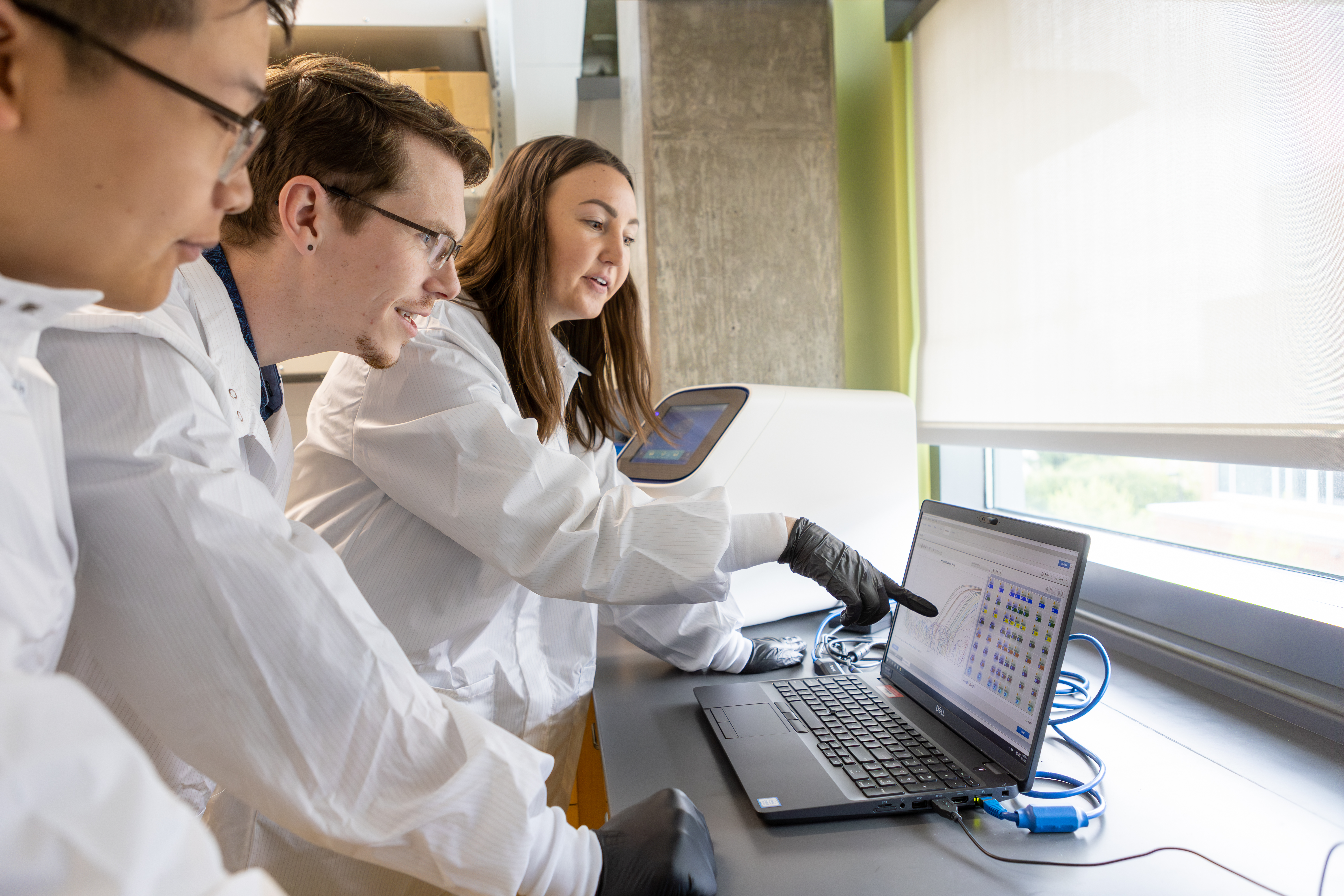Three Pacific University pharmacy students wearing lab coats look at a laptop in a lab