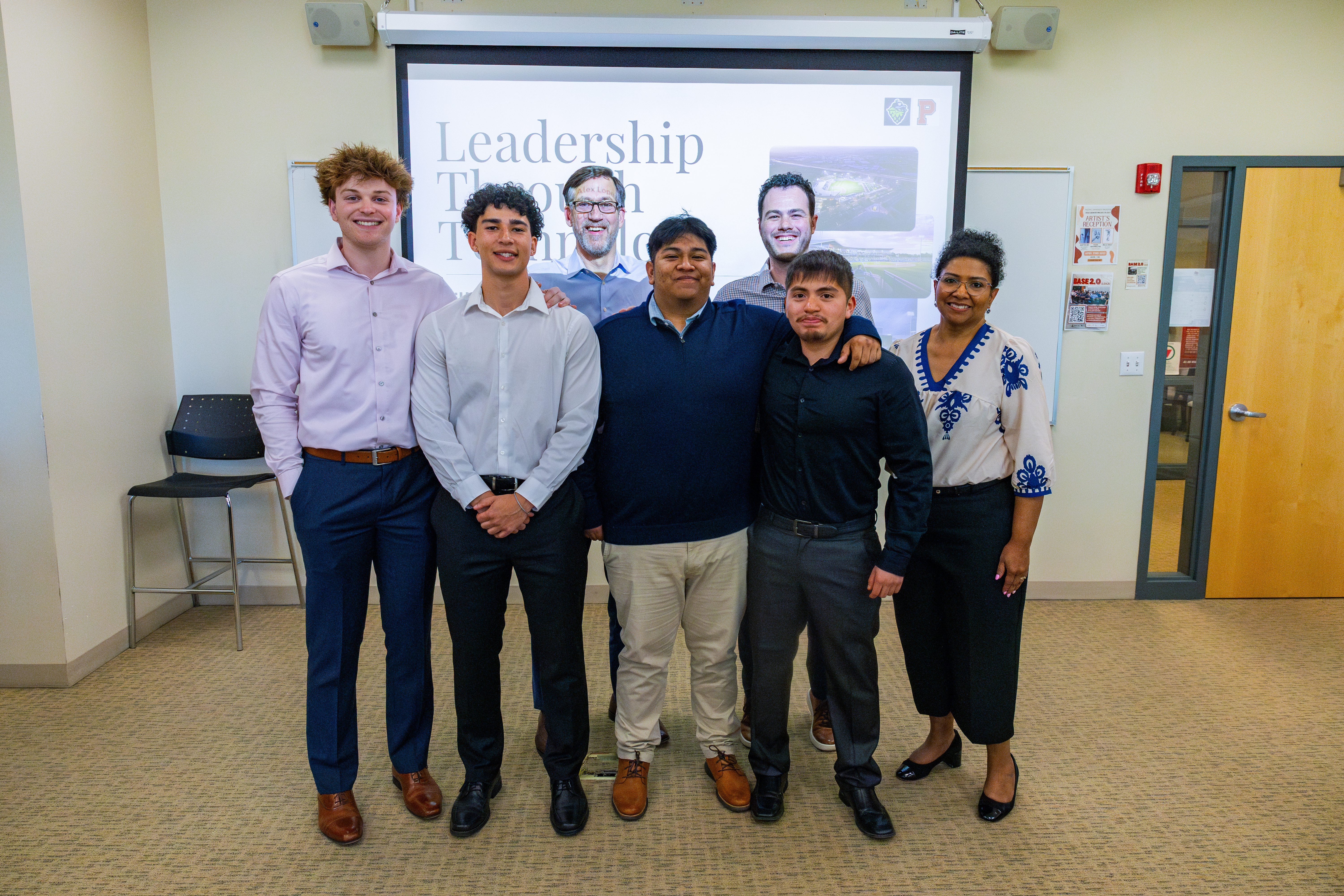 A group of Pacific University business students stand in a classroom in front of a presentation screen with the word "Leadership"