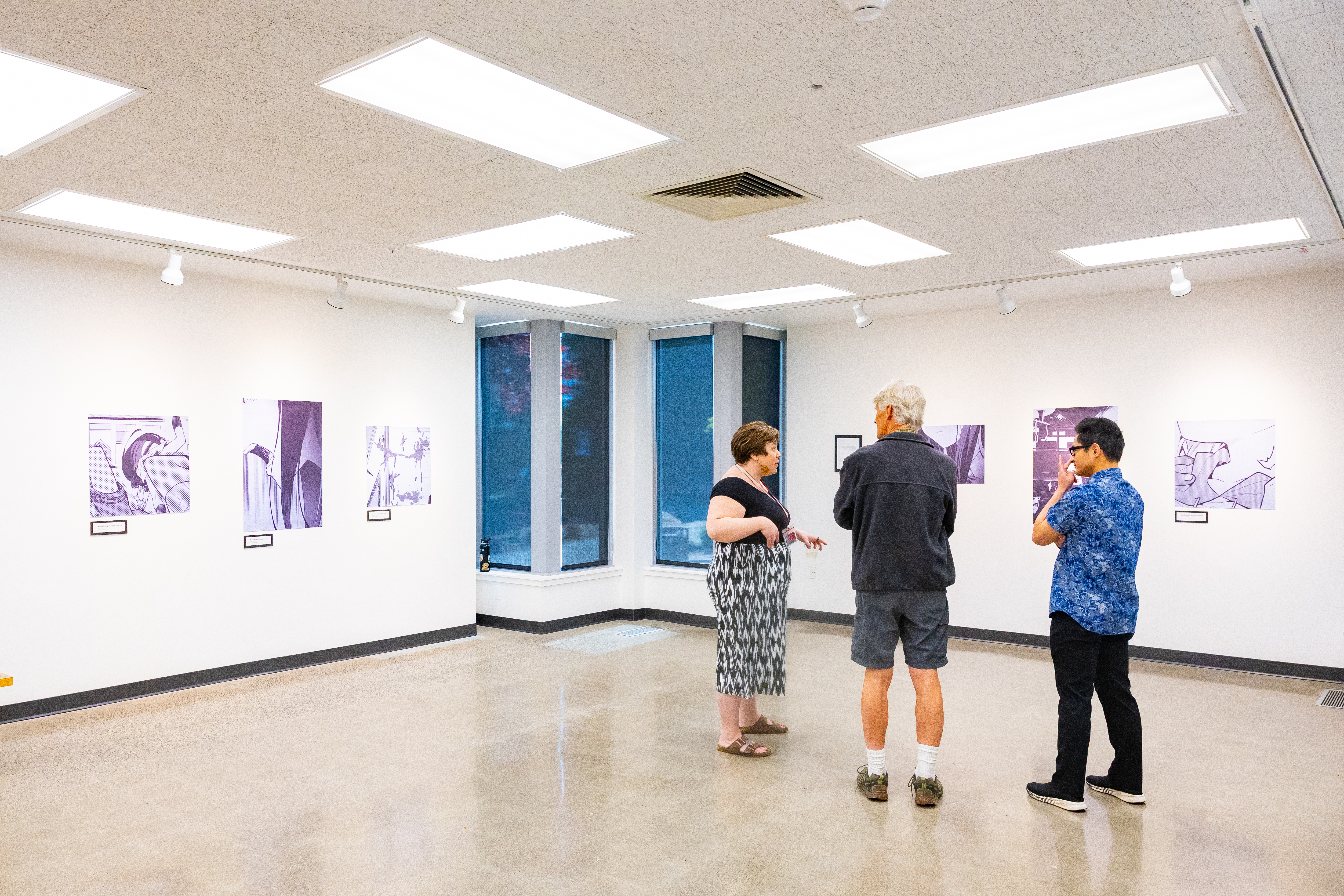 student at Pacific University exhibiting art work, six images hung on white walls, three people observing them