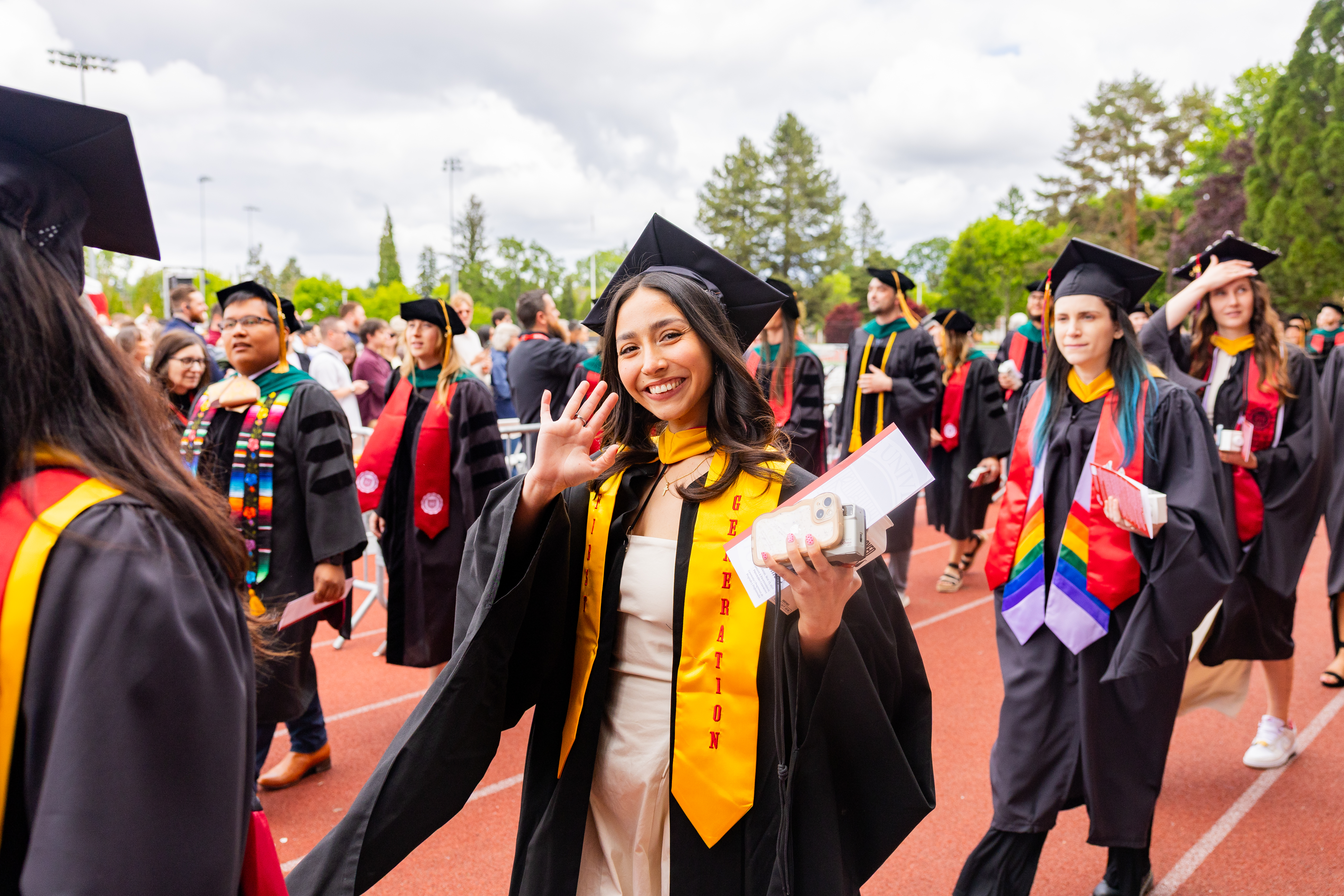 Pacific University students walking at commencement, one waving and wearing a first generation stole