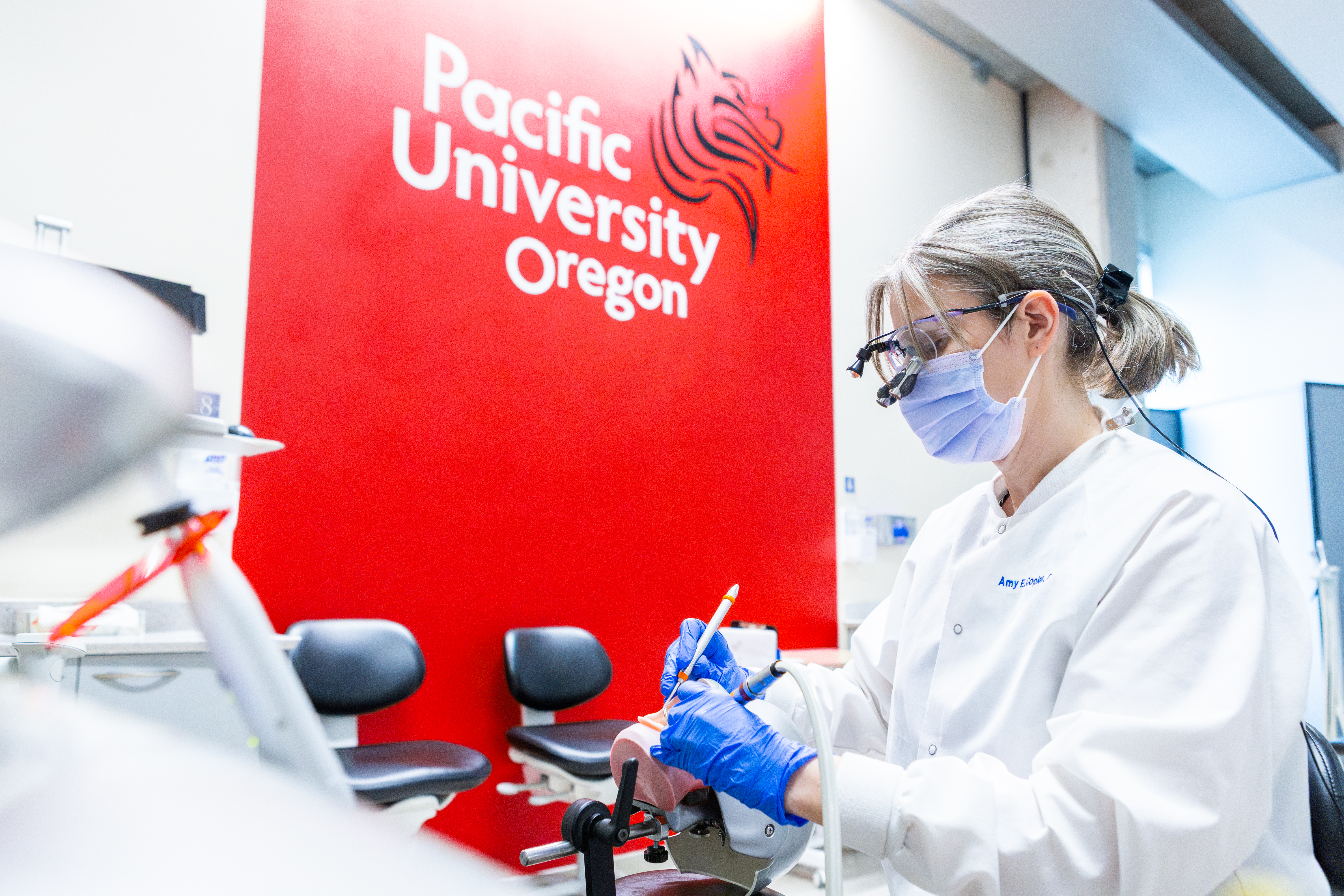 Amy Coplen, director of the school of dental hygiene studies, practices with a red Pacific University sign in background