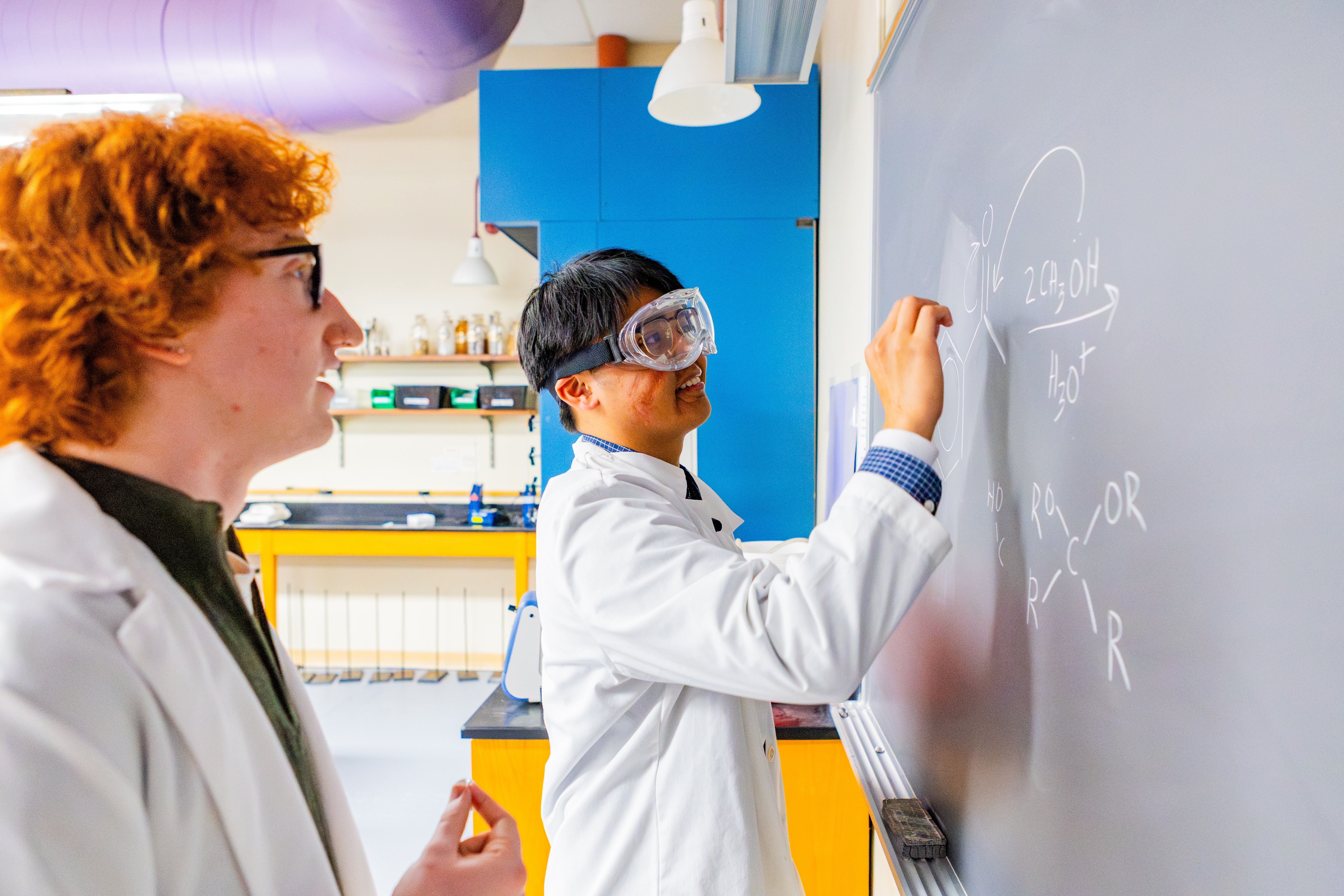 two Pacific University chemistry students write equations on a chalkboard while wearing lab coats