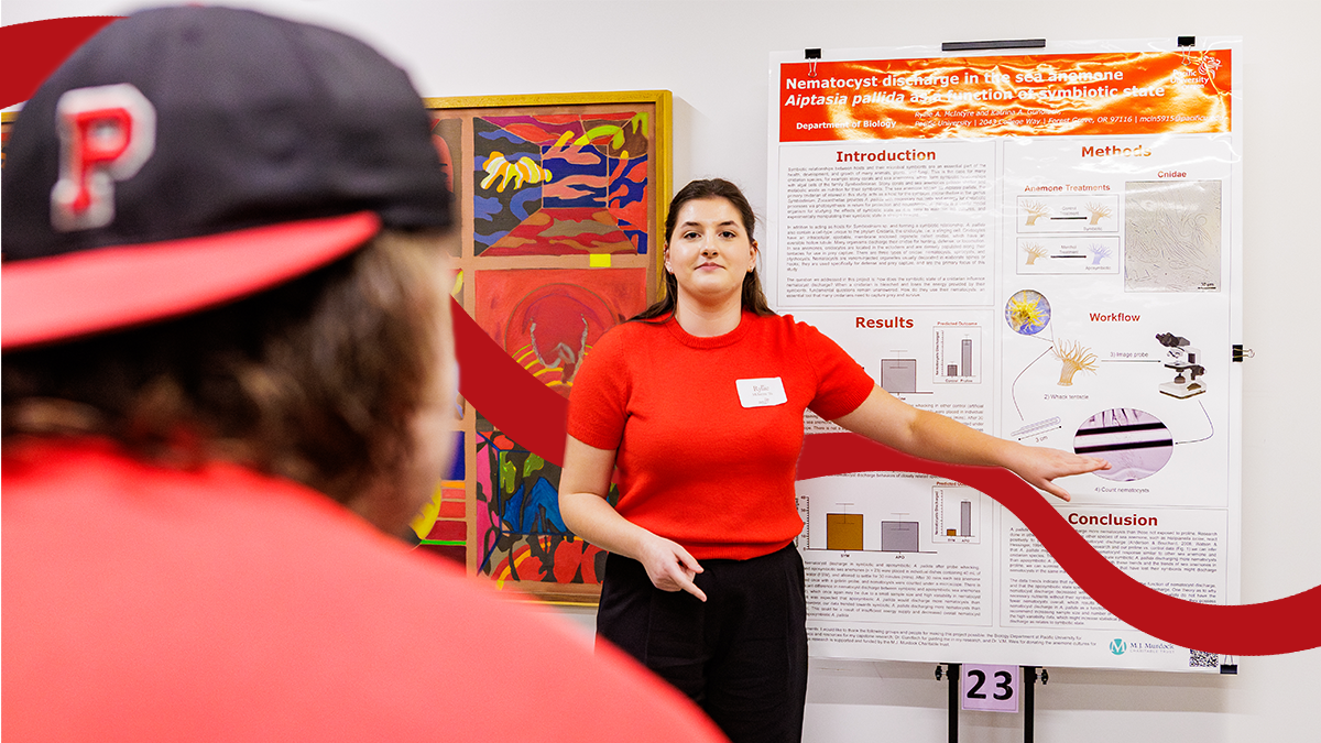 A Pacific student in a red shirt presents a research poster. In the foreground is an audience member wearing a backward Pacific hat.