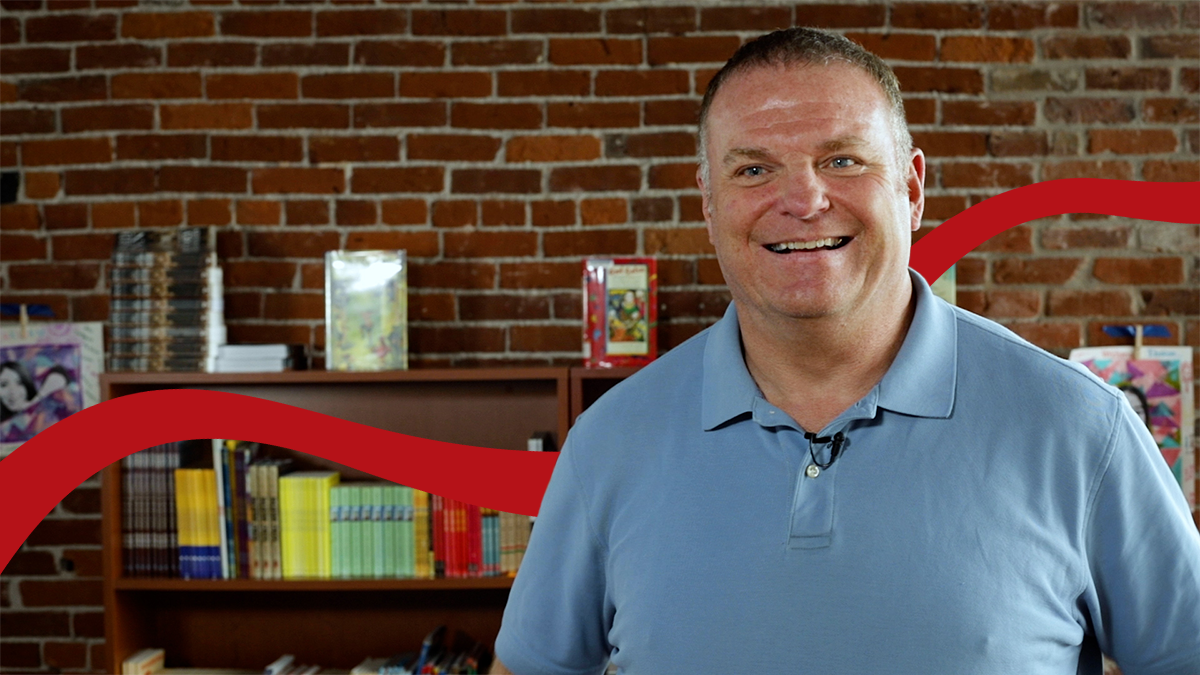 Kevin Carr, wearing a blue collared shirt, poses in front of a brick wall and a shelf full of books.