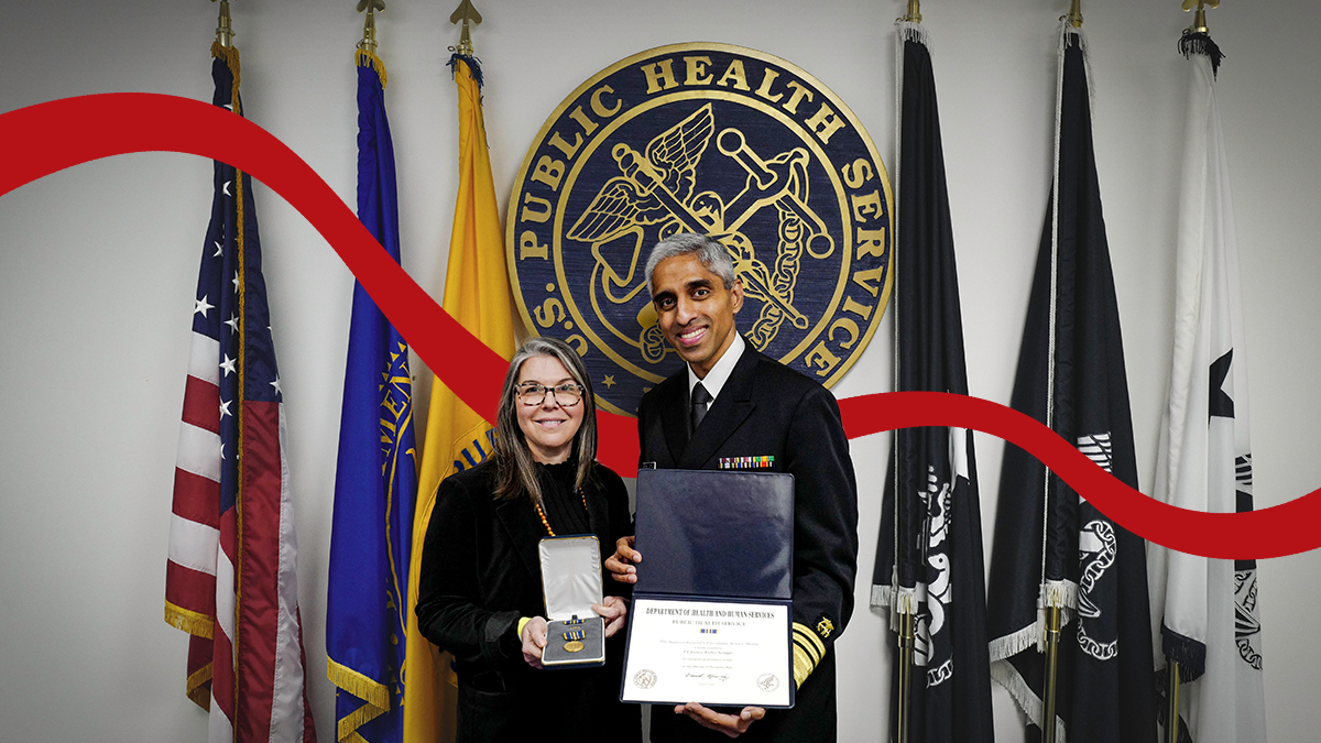 Jessica Scrugg '09 poses in front of a bank of flags with former U.S. Surgeon General Vitek Murthy. Murthy holds a certificate, and Scruggs holds a medal in a padded box.