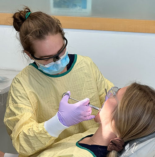 Alex Hays seated on left side in yellow gown, practicing giving a novocaine shot to another dental student.