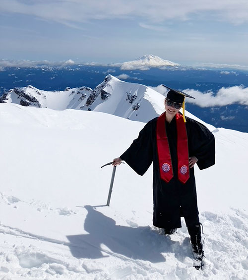 Alex Hays stands on top of a snow-covered Mount Saint Helens in her graduation gown and cap, with a Pacific University stoll on.