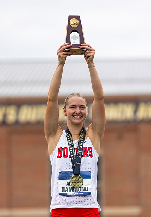 Woman with blonde hair. a white singlet and red shorts with a medal around her neck holds a trophy high above her head.