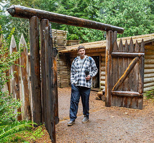Man with brown hair and in a plaid blakc and white overshirt stands inside the gate of Fort Clatsop.