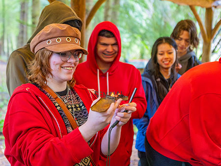 A student wearing a red shirt and brown hat with a burning tinderbox in her hands.