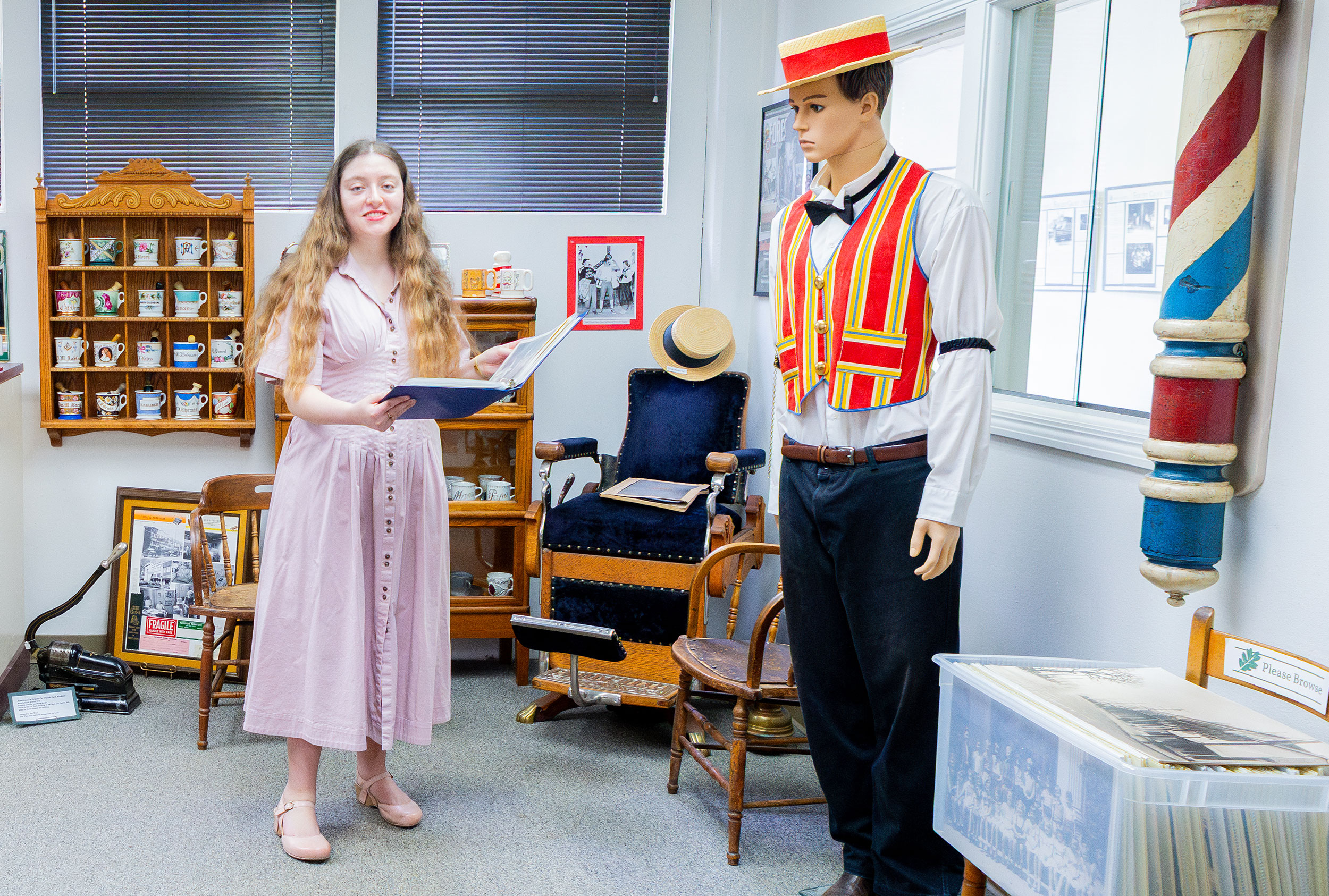 Elle Griego in a long dress, standing in a room with an antique barber's chair. a display with shaving mugs, and a mannequin wearing clothing from the 1890s.