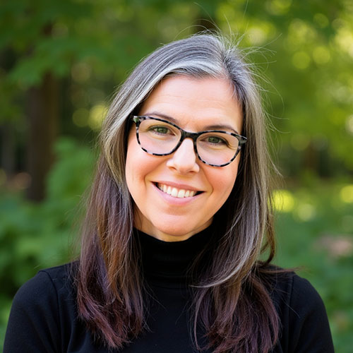Headshot of Jessica Scruggs DHS '09. She has long brown hair with highlights and is wearing a black turtleneck shirt and brown tortoise shell glasses. 