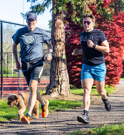 Two men running in blue shirts, one has a dog on a leash running alongside.