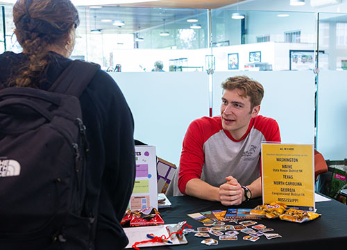 Kai Markle '27 in red and gray shirt talking with another student across a table. There is a yellow side describing voting registration deadlines.