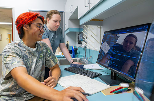 Two students review computer code or a large computer screen. The male student has dyed red hair, wear glasses and is wearing a camouflaged shirt. He holds a pen in his right hand, which hovers over the computer mouse. The female student has sandy brown hair pulled back into a bun and is wearing a slate blue shirt.