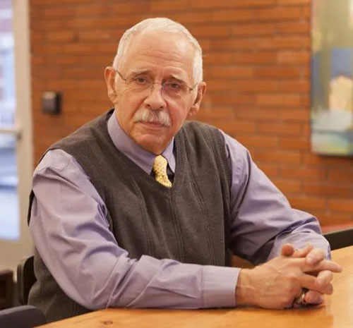 Mike Steele sits at a table. He has white hair and a white mustache and is wearing glasses. He is wearing a light blue dress shirt with a light yellow tie and a gray vest. His hands and folded in front of him.