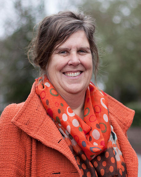 Headshot of Pacific Mathematics Professor Nancy Ann Neudauer. She has long brown hair, pulled up in a bun. She is wearing an orange jacket with wide lapels and an orange and brown scarf with red, gray and white rings printed on it.