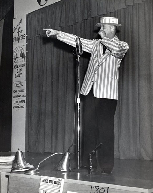 Main in a striped coat on stage points his hands past a microphone.