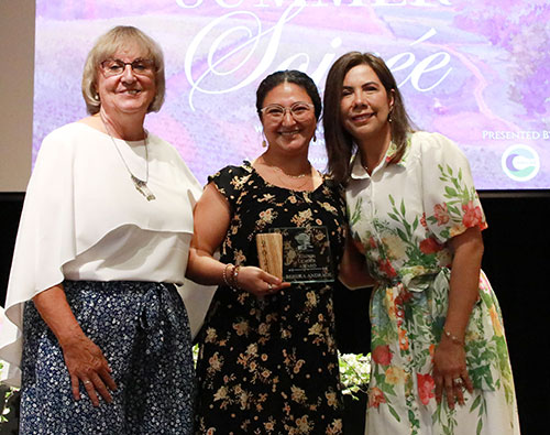 Three people presenting and receiving an award. All women, the one of the left is blonde, with glasses, wearing a white blouse ans a blue skirt. The person in the middle is Hispanic, wears glasses and is wearing a black dress with flowers. She is holding a wood and glass award. The woman on the right is Hispanic with long dark hair and is wearing a white dress with a floral print.