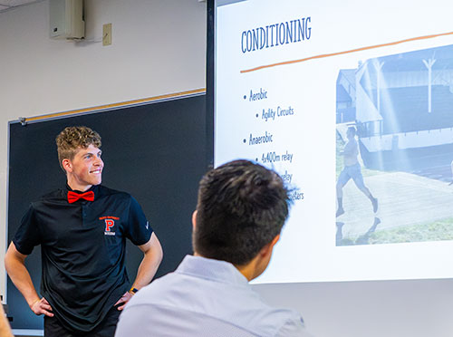 Man in black shirt and red bowtie with curly blonde hair presents in front of a classroom. The screen has a header of "Conditioning" with bullet points for a presentation.