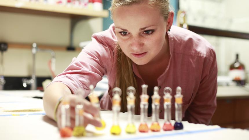 A female student aligns multicolored liquids in small glass jars by the darkness of their pigment.