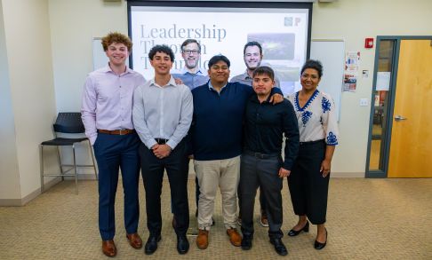 A group of Pacific University business students stand in a classroom in front of a presentation screen with the word "Leadership"