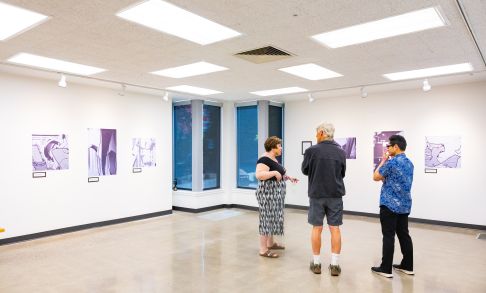 Pacific University student exhibiting art in a gallery setting, three people observe six pictures on a white wall