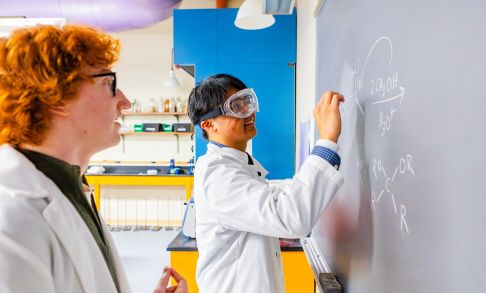 two Pacific University chemistry students write equations on a chalkboard while wearing lab coats
