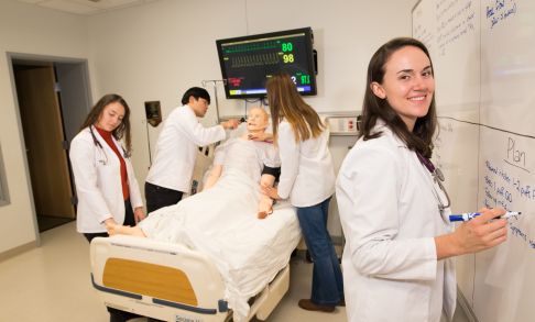 Four physician assistant program students demonstrate care on a dummy body lying in a hospital bed.