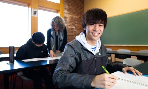 A student in a classroom looks into the camera between taking notes during a class.