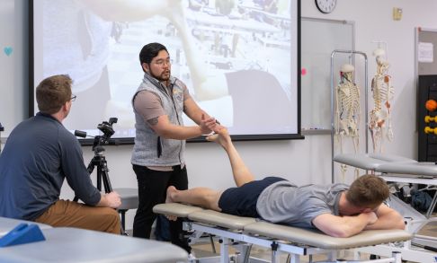 A physical therapy instructor demonstrates a way to strengthen the foot and leg on a student.