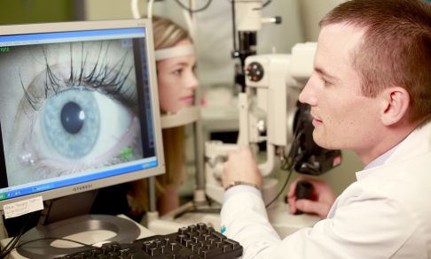 An optometry student examines a patient's eye using a large medical camera.