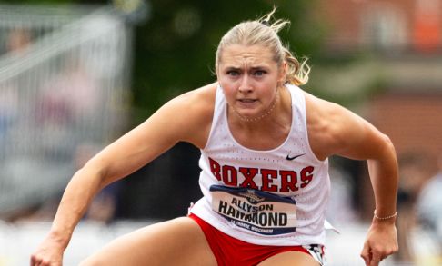 A women with blonde hair jumps over a hurdle. She has blonde hair and is wearing a white singlet with "BOXERS" in red across the front.