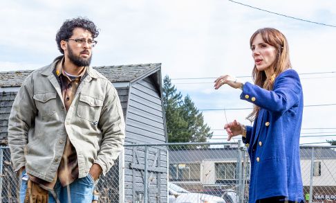 Two people, one a male with curly black hair and a beard, the other a woman in a blue blazer, discuss a topic outside in front of a gray house.