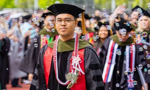 Jo Sean Ignaco PharmD '25 walks in academic regalia and hood with bubbles swirling in the air.