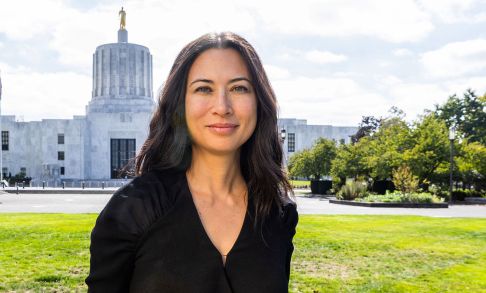 Rebecca Schoon stands in front of the Oregon State Capital. She is in the foreground, with blong black hair and wearing a black shirt. The white dome of the Oregon Capital building with the gold "Pioneer" statue is just behind her head.