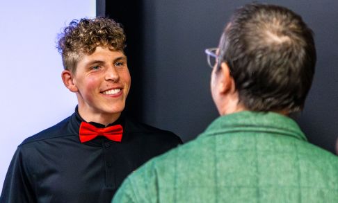 Tiger Reimann on left, in black shirt and red bow tie, talking with man in a green sweater with brown hair.