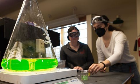 Chemistry Beaker With Yellow-Green Substances With Students Studying In Background