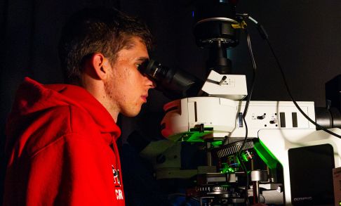 A man in a red hooded sweatshirt peers through a microcsope inside a dark lab room.
