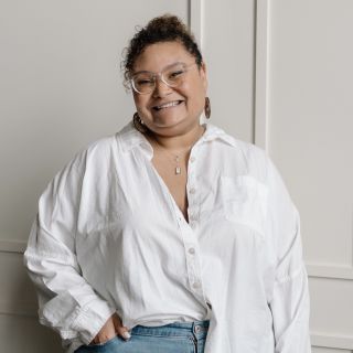 Black woman with curly hair in a bun, wearing glasses, earrings and a white button down shirt smiling at the camera.