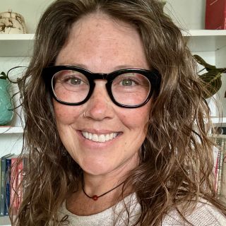 This is a headshot of Cristy Sellers. She is a white woman wearing a beige shirt, black rimmed glasses and she is standing in front of a white bookcase with plants, pottery, and books. She has wavy hair, freckles, and a nose ring. There is a necklace around her neck with a carnelian bead. 