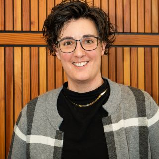 Dr. Mulcahy (a white queer person with black glasses and short curly dark hair, wearing a black turtleneck and grey/blue plaid sweater)) stands in front of a wooden background