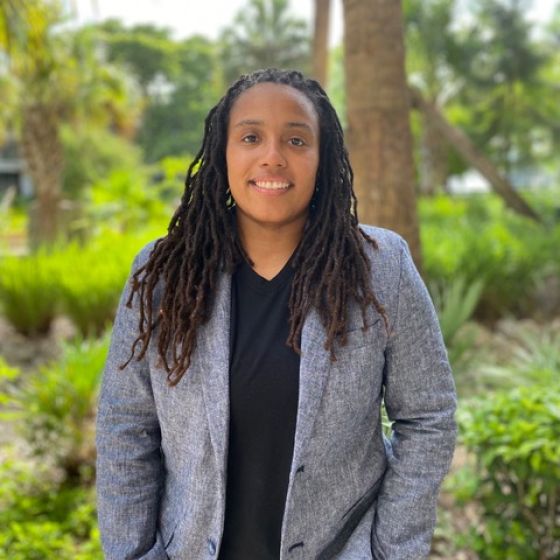 An image of Dr. Rochon outdoors. They are wearing a black shirt, gray blazer, and black pants. 