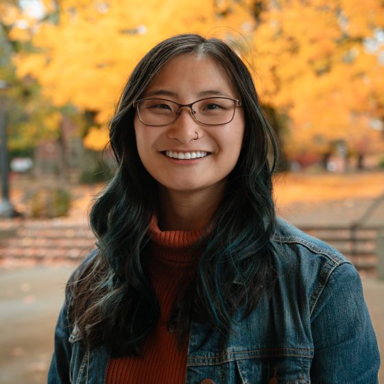 Headshot of Melissa Gabel, She Chinese. Her hair is dark with blue highlights. She is smiling and wearing a glasses and an orange turtleneck with a jean jack on top. The background is outdoors in front of Taylor-Meade