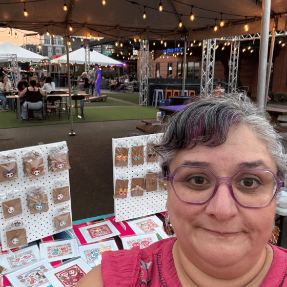 Headshot of a woman wearing glasses and a pink top smiling in an outdoor market setting.