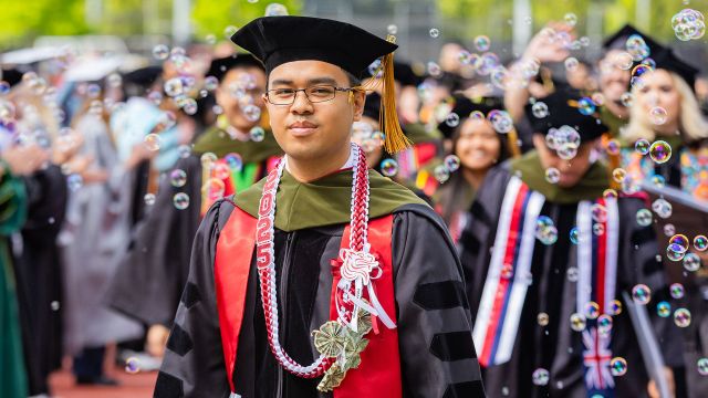 Jo Sean Ignaco PharmD '25 walks in academic regalia and hood with bubbles swirling in the air.